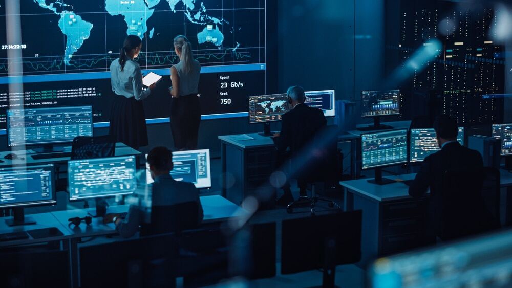 Two Software Engineers Talking Next to a Big Screen in a Modern Monitoring Office with Live Analysis Feed with Charts.